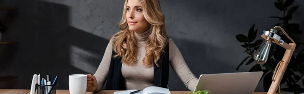 Panoramic shot of attractive and blonde businesswoman sitting at table and holding cup — Stock Photo