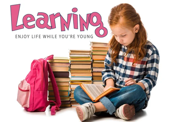 Positive redhead schoolkid sitting near books and learning enjoy life while you are young letters on white — Stock Photo