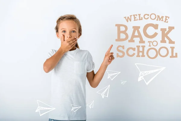 Enfant choqué couvrant visage tout en regardant la caméra et pointant du doigt à bienvenue lettres de retour à l'école sur blanc — Photo de stock