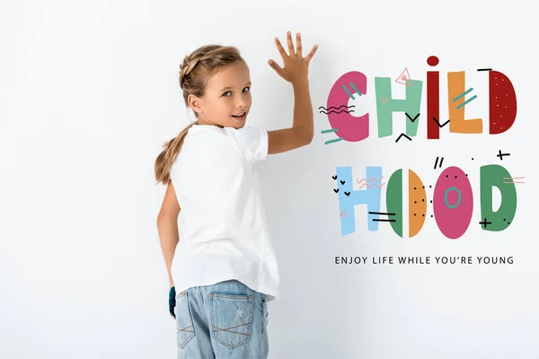 Happy kid looking at camera near childhood enjoy life while you are young letters on white — Stock Photo