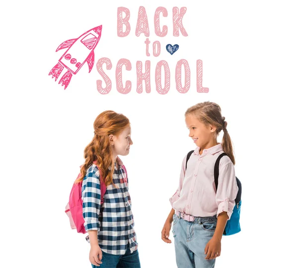 Niños felices con mochilas mirándose el uno al otro cerca de las letras de la escuela en blanco - foto de stock