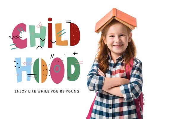 Cheerful redhead pupil with orange book on head standing with crossed arms near childhood enjoy life while you are young letters on white — Stock Photo