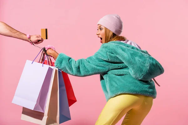 Cropped view of male hand with credit card handcuffed with aggressive african american woman holding shopping bags on pink background, fashion doll concept — Stock Photo