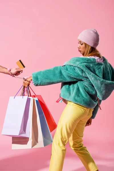 Cropped view of male hand with credit card handcuffed with offended african american woman holding shopping bags on pink background, fashion doll concept — Stock Photo