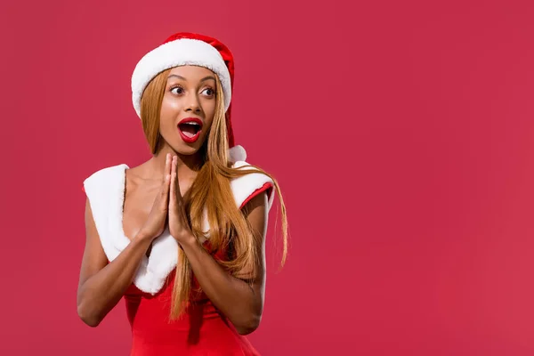 Amüsiertes afrikanisch-amerikanisches Mädchen mit Weihnachtsmütze und Weihnachtskleid, das mit gefalteten Händen steht und isoliert auf rotem Grund wegschaut — Stockfoto