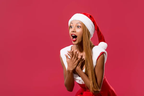 Surprised african american girl in santa hat and christmas dress looking away isolated on red — Stock Photo