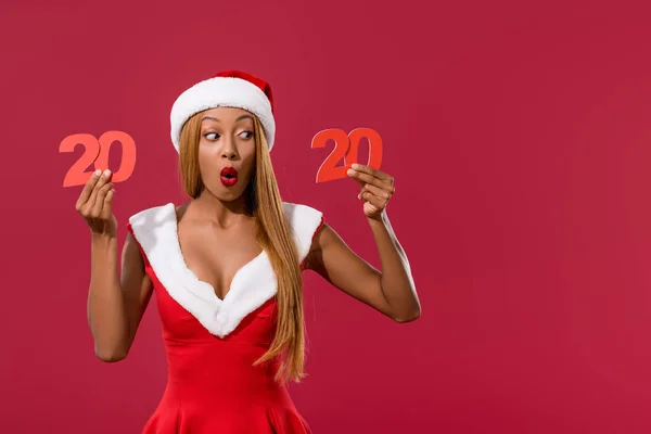 Surprised african american girl in santa hat and christmas dress holding 2020 paper cut number isolated on red — Stock Photo