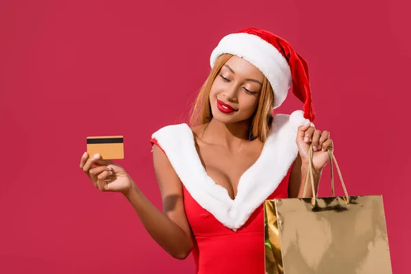 Niña afroamericana sonriente en sombrero de santa y vestido de navidad con tarjeta de crédito y bolsa aislada en rojo - foto de stock