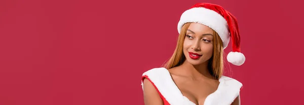 Colpo panoramico di ragazza afro-americana in cappello di Babbo Natale e vestito di Natale guardando lontano isolato sul rosso — Foto stock