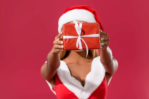 Giovane ragazza afroamericana in cappello di Babbo Natale e vestito di Natale in possesso di scatola regalo di fronte al viso isolato sul rosso — Foto stock