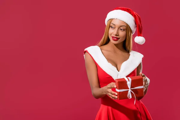 Sexy african american girl in santa hat and christmas dress holding gift box isolated on red — Stock Photo