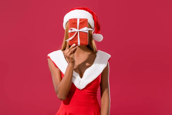 Young african american girl in santa hat and christmas dress hiding face behind gift box isolated on red — Stock Photo