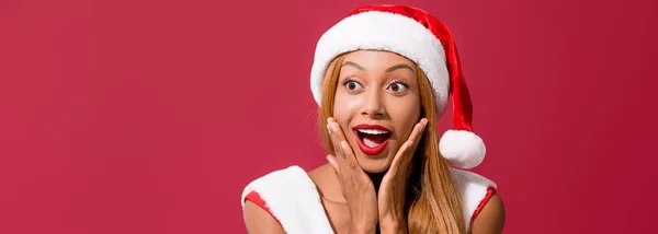 Panoramic shot of african american girl in santa hat holding hands near face isolated on red — Stock Photo