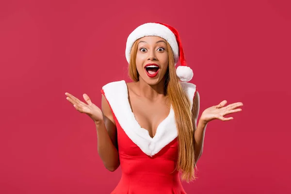 Amused aftrican american girl in santa hat and christmas dress showing wow gesture isolated on red — Stock Photo