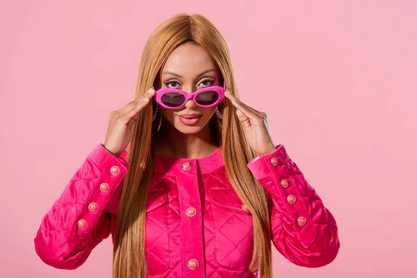Elegante donna afroamericana toccando occhiali da sole e guardando la fotocamera isolata sul rosa, concetto di bambola di moda — Foto stock