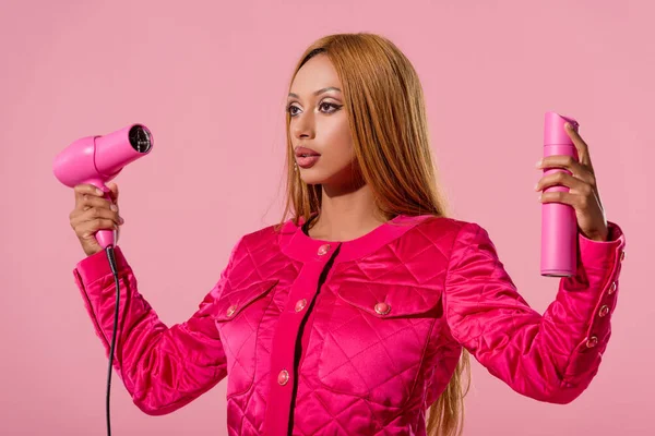 Hermosa, elegante mujer afroamericana sosteniendo spray para el cabello y secador de pelo aislado en rosa, concepto de muñeca de moda - foto de stock