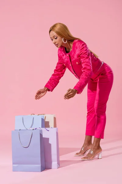 Mujer afroamericana de moda mirando bolsas de compras sobre fondo rosa, concepto de muñeca de moda - foto de stock
