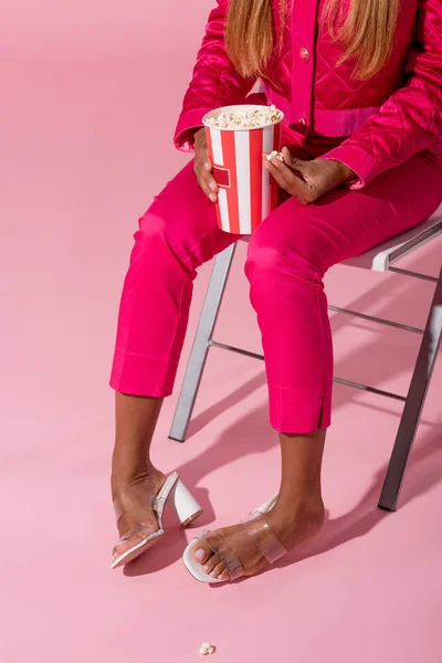 Corte vista de stylsh Africano americano mulher sentada na cadeira com xícara de pipoca no fundo rosa, conceito de boneca de moda — Fotografia de Stock