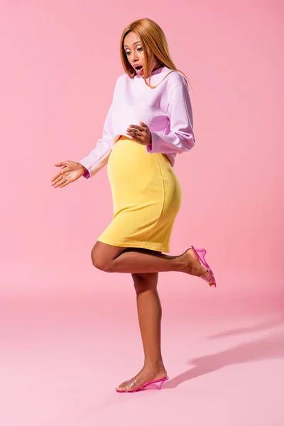 Shocked, pregnant african american woman standing on one leg on pink background, fashion doll concept — Stock Photo