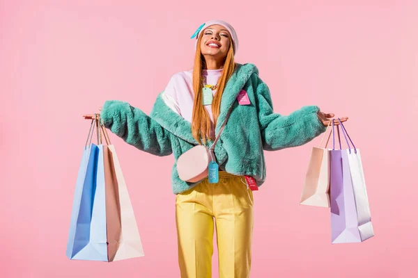 Felice, donna afro-americana elegante in piedi con gli occhi chiusi e tenendo borse della spesa isolate su rosa, concetto di bambola di moda — Foto stock