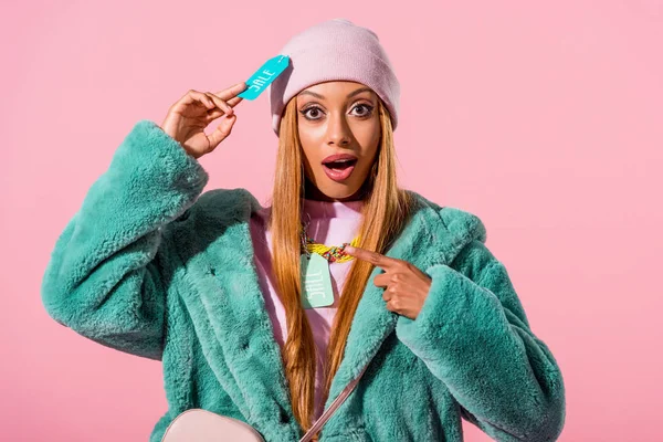 Amüsiert, stilvolle afrikanisch-amerikanische Frau anrührendes Label mit Verkaufsaufschrift und Zeigefinger isoliert auf rosa, Modepuppenkonzept — Stockfoto