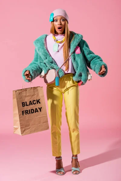 Chocado, elegante menina africana ameican segurando saco de compras com inscrição sexta-feira preta no fundo rosa, conceito de boneca de moda — Fotografia de Stock