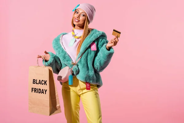 Весела, модна африканська дівчина тримає кредитну картку і сумку з чорним святим написом ізольована на рожевій, концепції модної ляльки — стокове фото