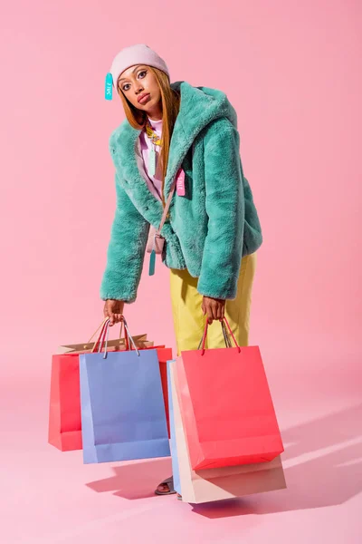 Exhausted, fashionable african american woman holding shopping bags on pink background, fashion doll concept — Stock Photo