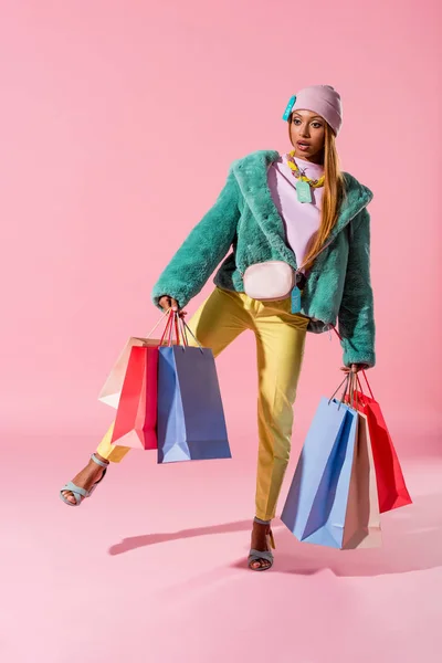 Atraente, mulher americana africana elegante segurando sacos de compras enquanto está em uma perna no fundo rosa, conceito de boneca de moda — Fotografia de Stock