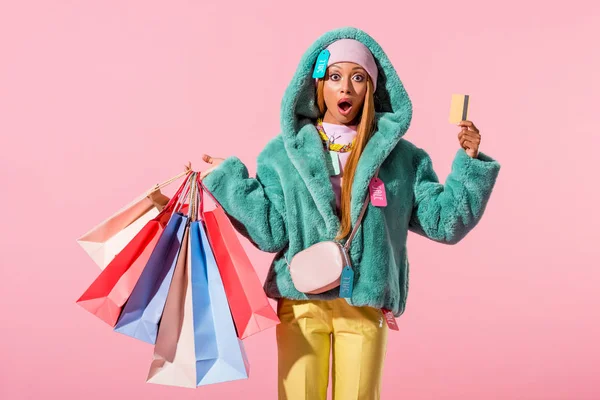 Surprised, stylish african american woman holding credit card and shopping bags on pink background, fashion doll concept — Stock Photo