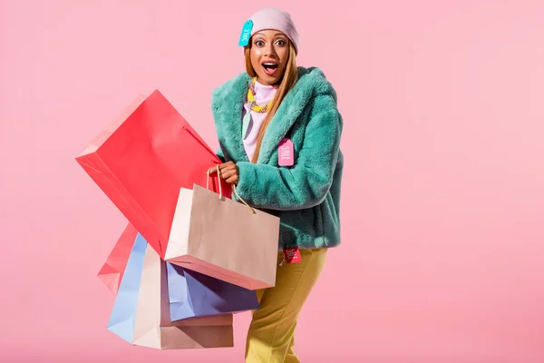Emocionado, mujer afroamericana con estilo sosteniendo bolsas de compras y mirando a la cámara aislada en rosa, concepto de muñeca de moda - foto de stock