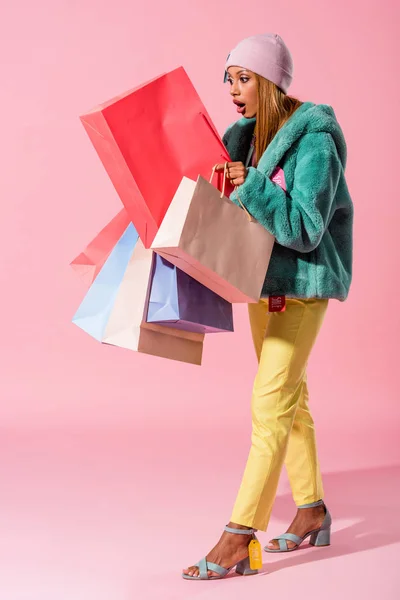 Surprised, trendy african american woman looking in shopping bag on pink background, fashion doll concept — Stock Photo