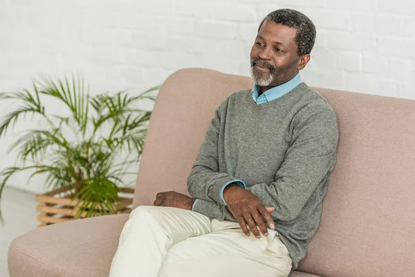 Мрійливий, усміхнений афроамериканський чоловік сидить на дивані вдома — стокове фото