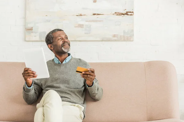Hombre afroamericano mayor sosteniendo tableta digital y tarjeta de crédito mientras está sentado en el sofá y mirando hacia otro lado - foto de stock