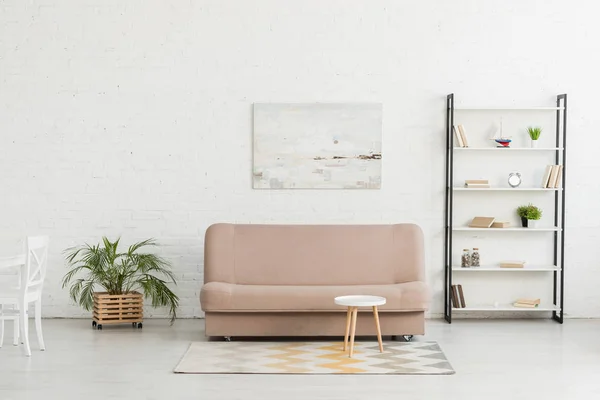 Espaçosa sala de estar com parede branca, sofá, rack com livros, mesa redonda e vasos de plantas — Fotografia de Stock