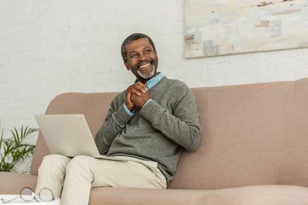 Feliz homem americano africano sentado no sofá com laptop e olhando para longe — Fotografia de Stock
