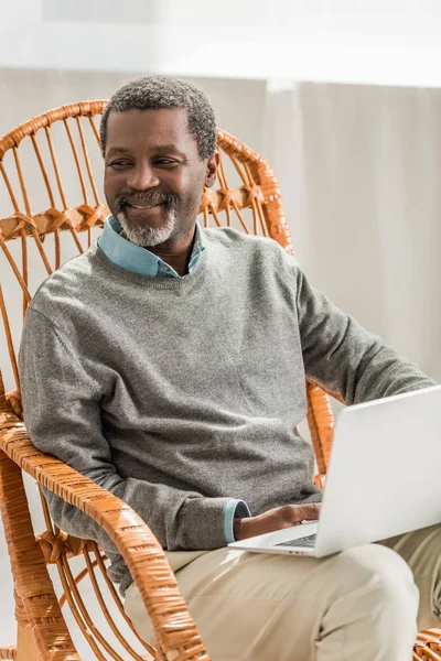 Sonriente afroamericano hombre sentado en silla de mimbre con portátil - foto de stock