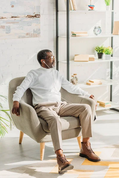 Старший африканский американец в стильной одежде сидит в кресле и слушает музыку в наушниках — стоковое фото