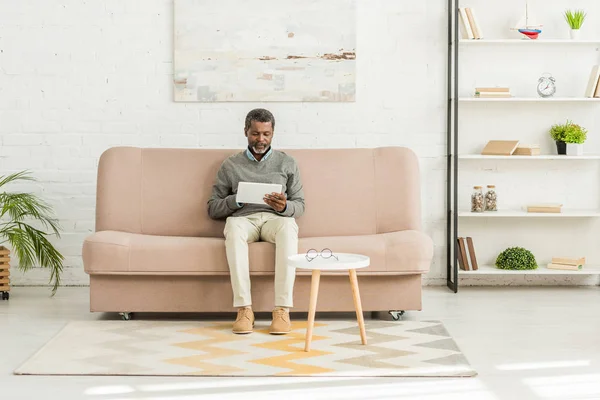 Homem americano africano sênior sentado no sofá na sala de estar e usando tablet digital — Fotografia de Stock