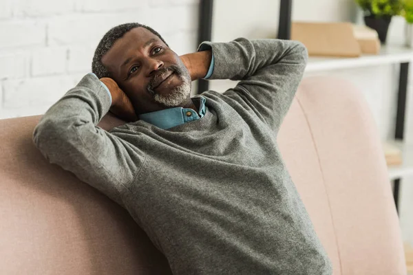 Hombre afroamericano soñador y positivo mirando hacia otro lado mientras está sentado en el sofá - foto de stock