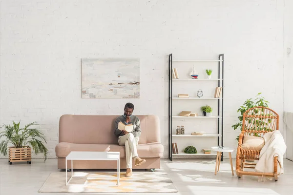 Homme afro-américain senior livre de lecture tout en étant assis sur le canapé dans le salon — Photo de stock