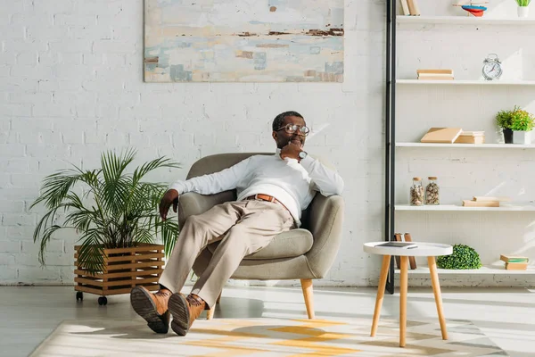 Hombre afroamericano elegante descansando en sillón y mirando hacia otro lado - foto de stock