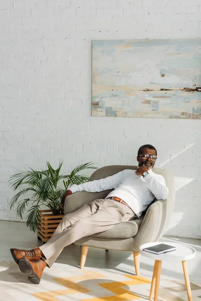 Hombre afroamericano elegante descansando en sillón y mirando hacia otro lado - foto de stock