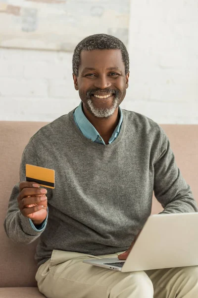 Старший африканский американец сидит на диване с ноутбуком, держит кредитку и улыбается в камеру — стоковое фото
