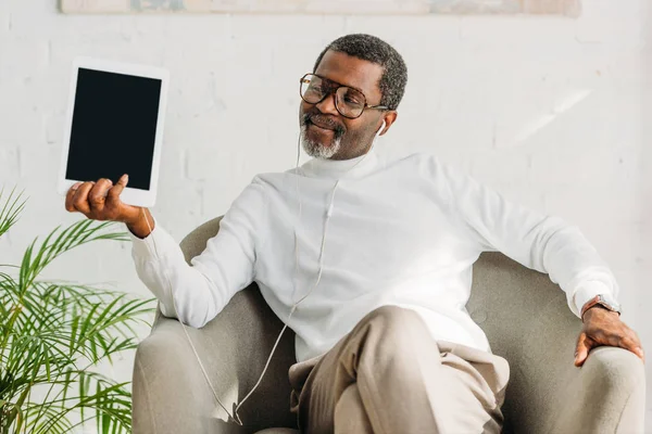 Hombre afroamericano elegante sentado en sillón, escuchando música en auriculares y sosteniendo la tableta digital - foto de stock