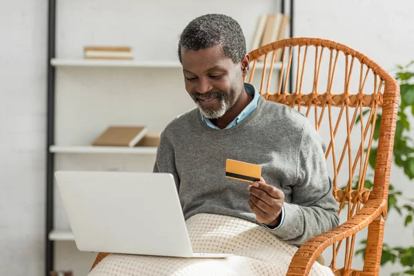 Uomo afroamericano sorridente seduto in poltrona di vimini con computer portatile e in possesso di carta di credito — Foto stock