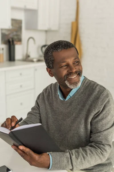 Anziano uomo afroamericano sorridente e distogliendo lo sguardo mentre scrive nel taccuino — Foto stock
