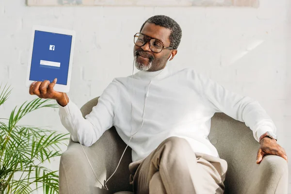 KYIV, UCRANIA - 2 de octubre de 2019: Elegante hombre afroamericano con auriculares que muestra la tableta digital con la aplicación de Facebook en la pantalla . - foto de stock