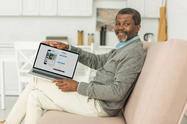 Kyiv, ukraine - 2. Oktober 2019: fröhlicher afrikanisch-amerikanischer Mann sitzt auf Sofa und zeigt Laptop mit Instagram-App auf dem Bildschirm. — Stockfoto