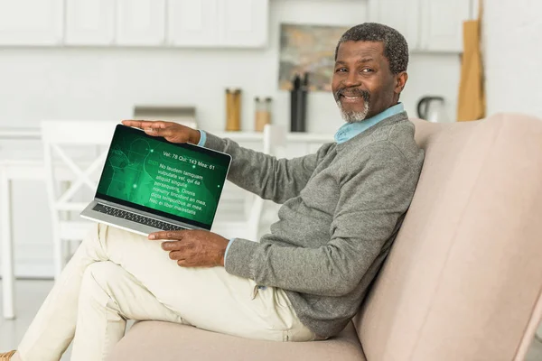 КИЕВ, УКРАИНА - 2 ОКТЯБРЯ 2019 года: Веселый африканский американец сидит на диване и показывает ноутбук с медицинской информацией на экране . — стоковое фото